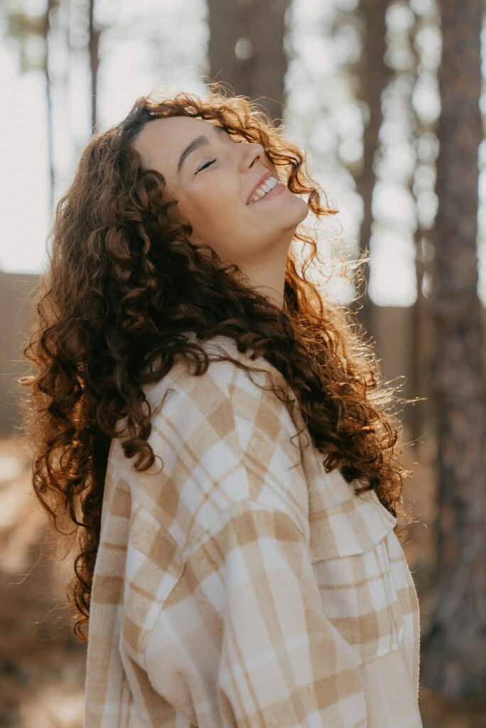pexels-photo-26214064-26214064 Smiling woman enjoying nature with eyes closed, curly hair shimmering in sunlight.