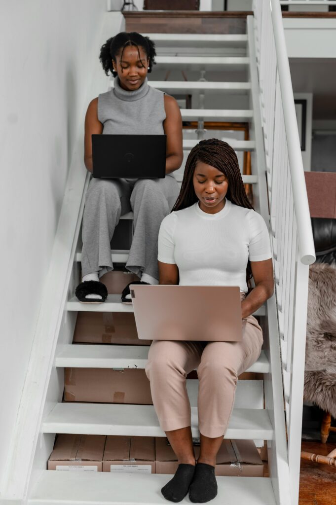 pexels-photo-8091336-8091336 Two women working remotely on laptops from home sitting on stairs indoors.